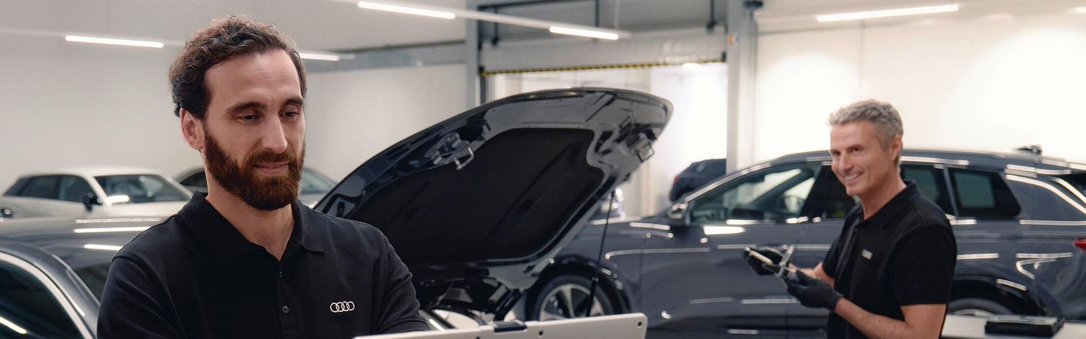 Zwei Audi Service Techniker in einer Werkstatt unterhalten sich. Einer von beiden blickt dabei auf seinen Laptop.