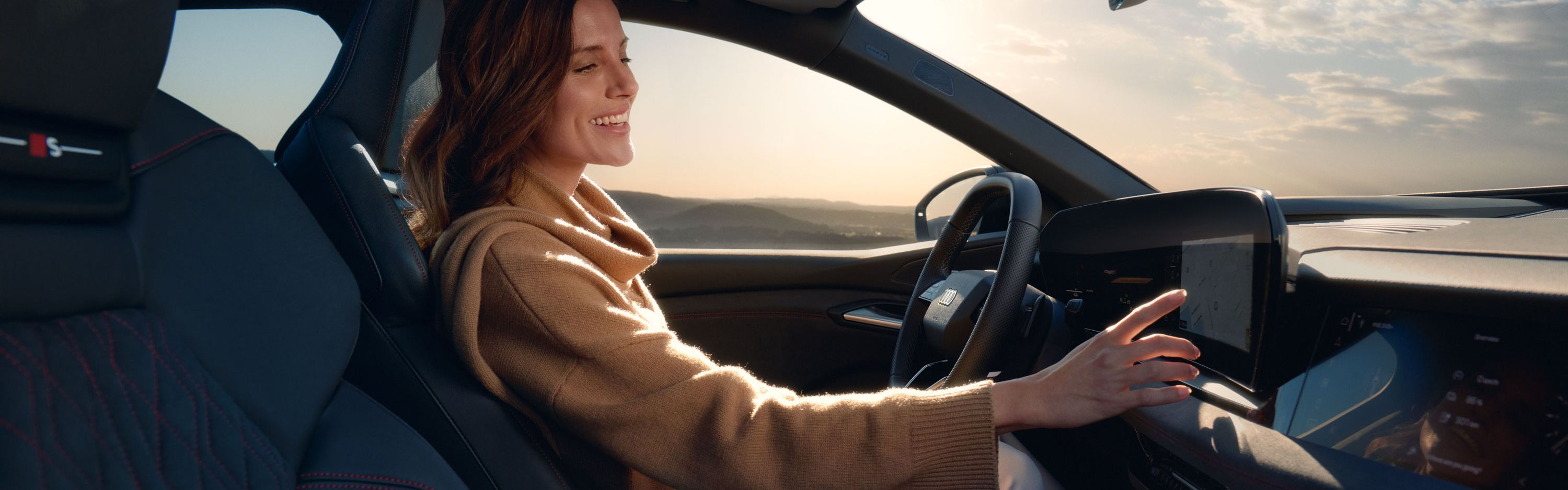 Junge Frau sitzt lächelnd am Steuer eines Audi und bedient den Touchscreen des MMI. Im Hintergrund ist eine weite Landschaft unter leicht bewölktem Himmel zu sehen.