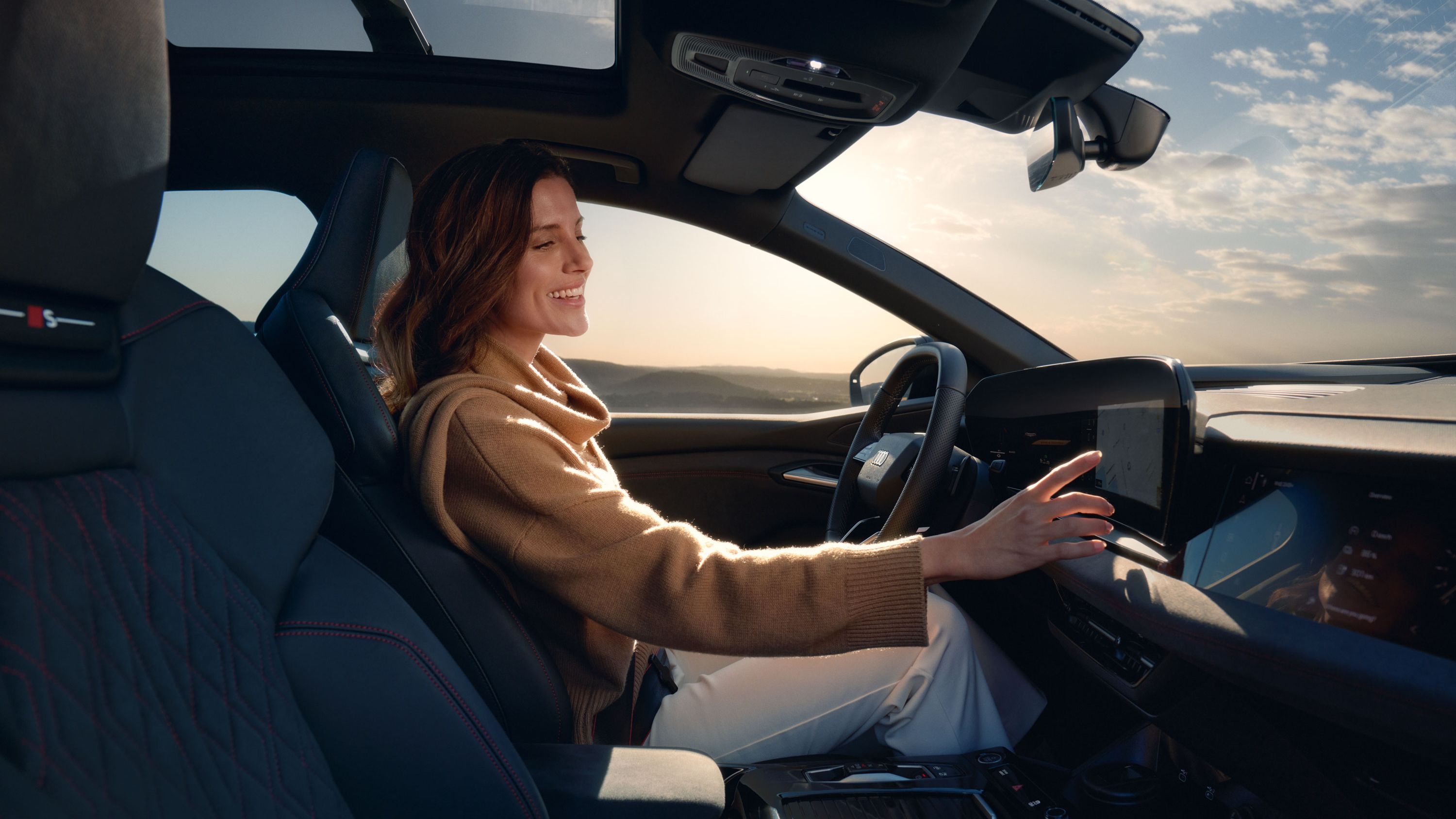 Junge Frau sitzt lächelnd am Steuer eines Audi und bedient den Touchscreen des MMI. Im Hintergrund ist eine weite Landschaft unter leicht bewölktem Himmel zu sehen.