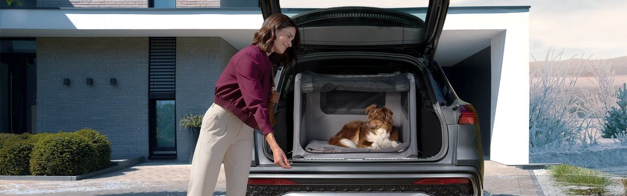 Eine Frau öffnet den Kofferraum eines geparkten Audi SUVs vor einem modernen Haus und streckt die Hand zu ihrem Hund aus, der entspannt im Laderaum liegt. Die Einfahrt ist leicht mit Schnee bedeckt, während der Himmel klar und winterlich wirkt.