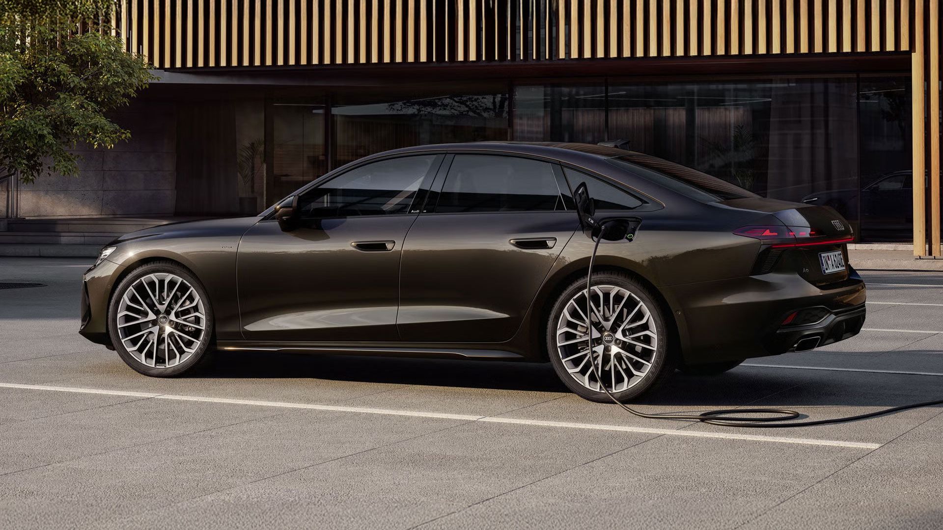 Die A6 Limousine e-hybrid in Havannaschwarz Metallic steht seitlich vor einer Gebäudefassade und ist dabei an eine Ladestation angeschlossen.