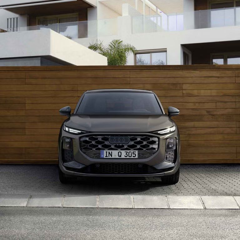 Der Q3 Sportback in Frontansicht parkt vor einem modernen Gebäude, dahinter eine Wand in Holzoptik.