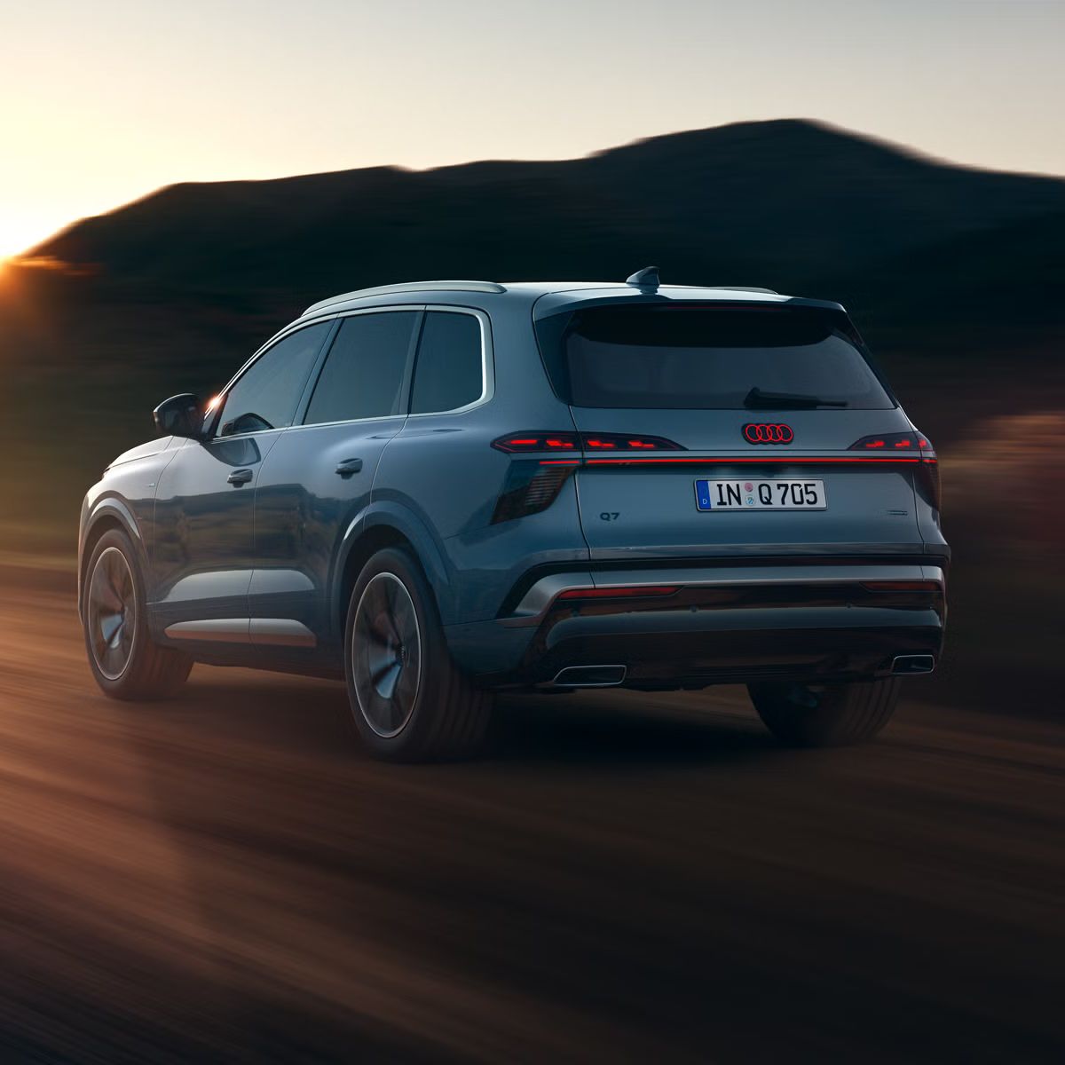 Der Audi Q7 SUV schräg von hinten in voller Fahrt auf einer Landstraße, im Hintergrund Gebirge. Die am Horizont untergehende Sonne beleuchtet die Szenerie.  