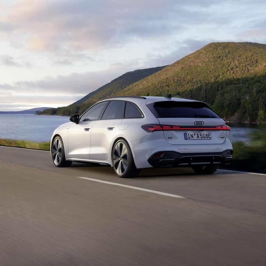 Der A5 Avant e-hybrid in einer schrägen Heckperspektive, während er eine Straße am Ufer eines Sees entlangfährt.