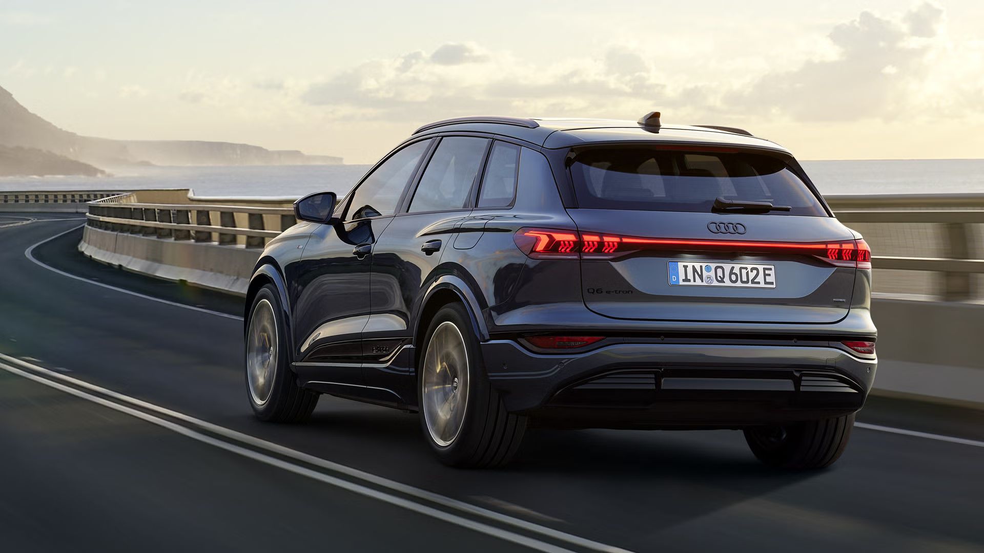 Schräge Heckaufnahme des Q6 SUV e-tron, welche über eine kurvige Straße am Meer fährt. Im Hintergrund sind Berge zu sehen.