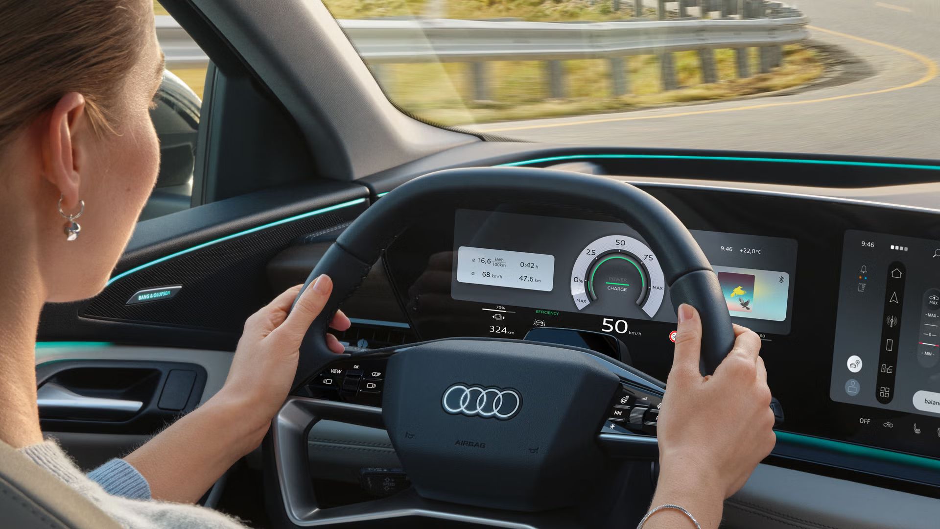 Cockpit des Q6 SUV e-tron aus Rücksitzperspektive. Eine junge Frau sitzt auf dem Fahrersitz und hält das Lenkrad mit beiden Händen fest. Durch die Windschutzscheibe sieht man eine Straße in der Natur.