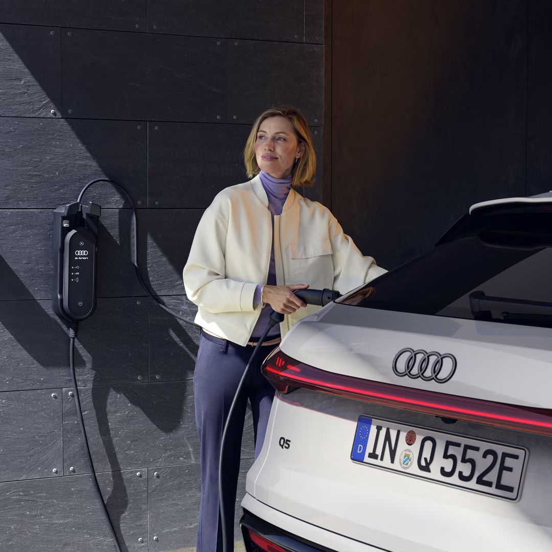 Blick auf das Heck des Q5 Sportback e-hybrid. Eine junge Frau schließt das Fahrzeug an eine Ladestation an.