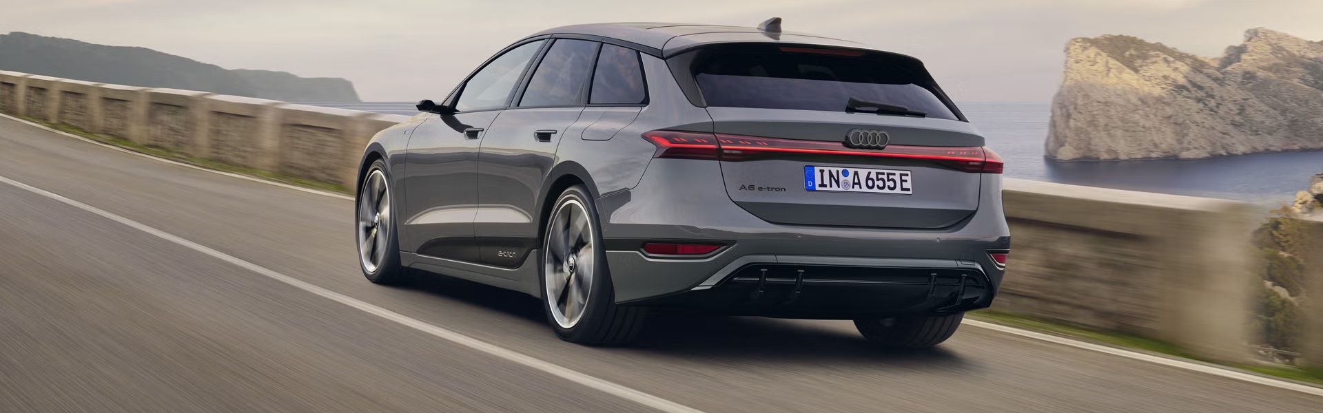 Schräge Heckaufnahme des A6 Avant e-tron, welcher auf einer Straße entlang des Meeres fährt. Im Hintergrund sind Felsen und Berge zu erkennen. 