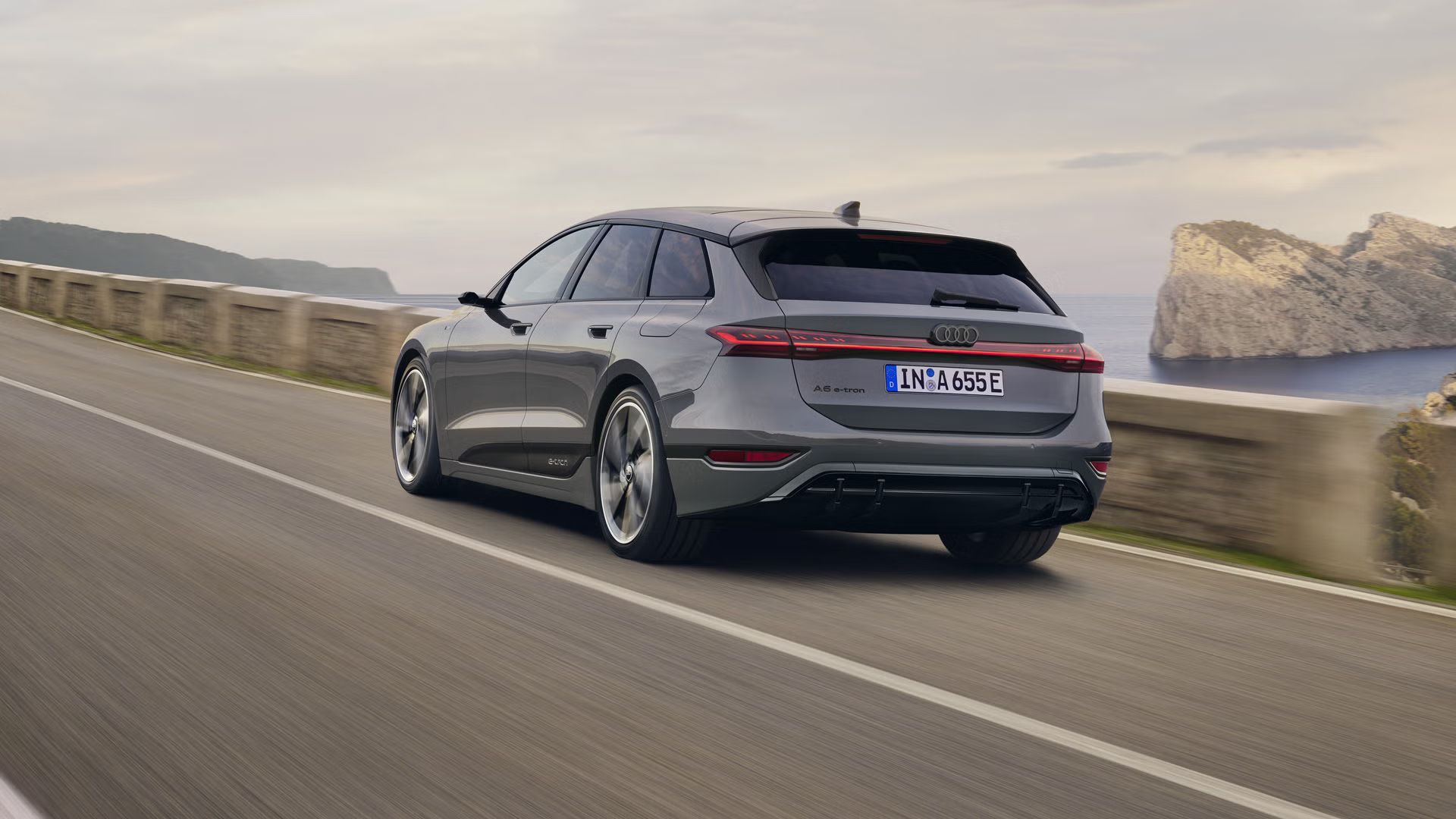 Schräge Heckaufnahme des A6 Avant e-tron, welcher auf einer Straße entlang des Meeres fährt. Im Hintergrund sind Felsen und Berge zu erkennen. 