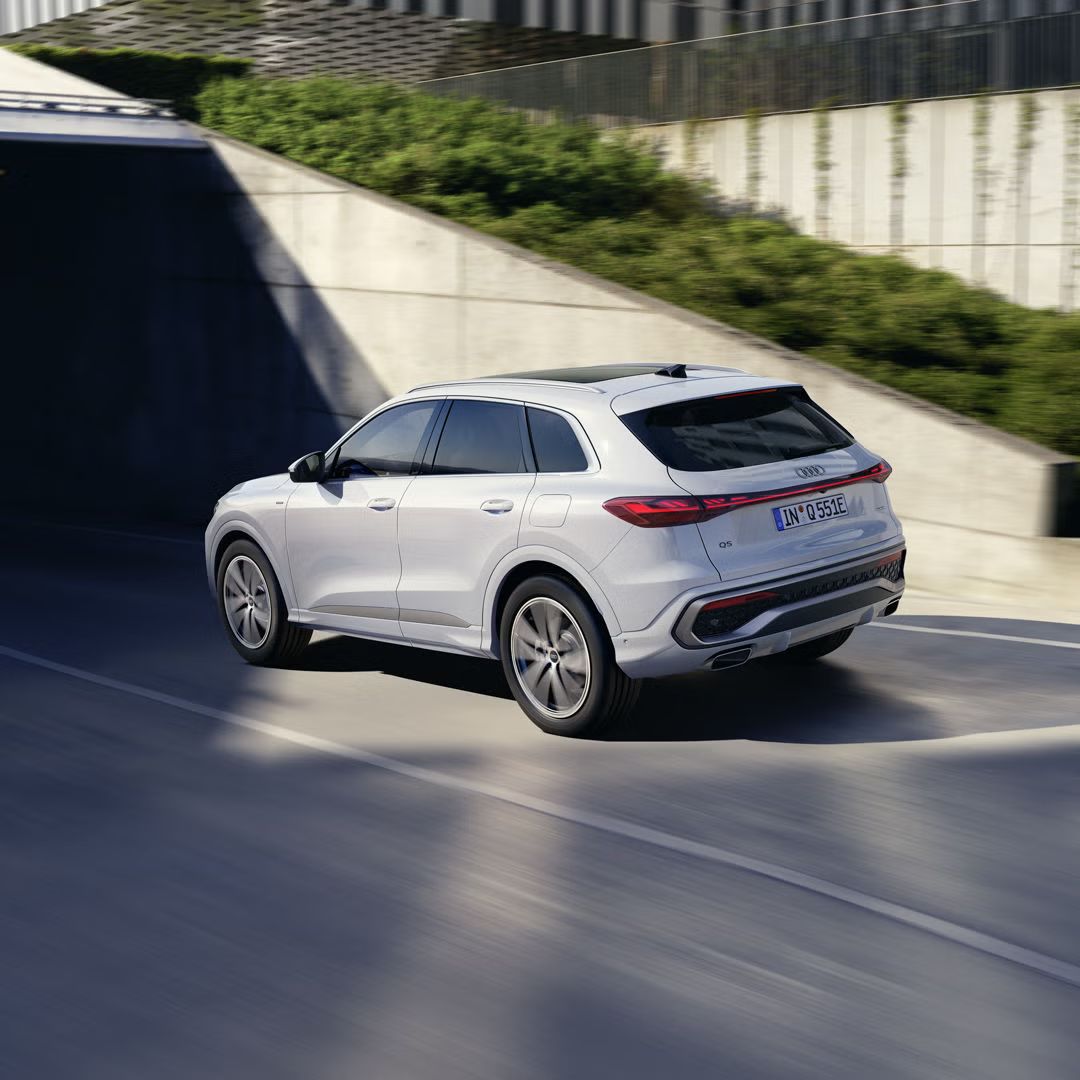 Schräge, hintere Ansicht auf einen weißen Q5 SUV e-hybrid, welcher auf einer Straße in einer Stadt fährt.