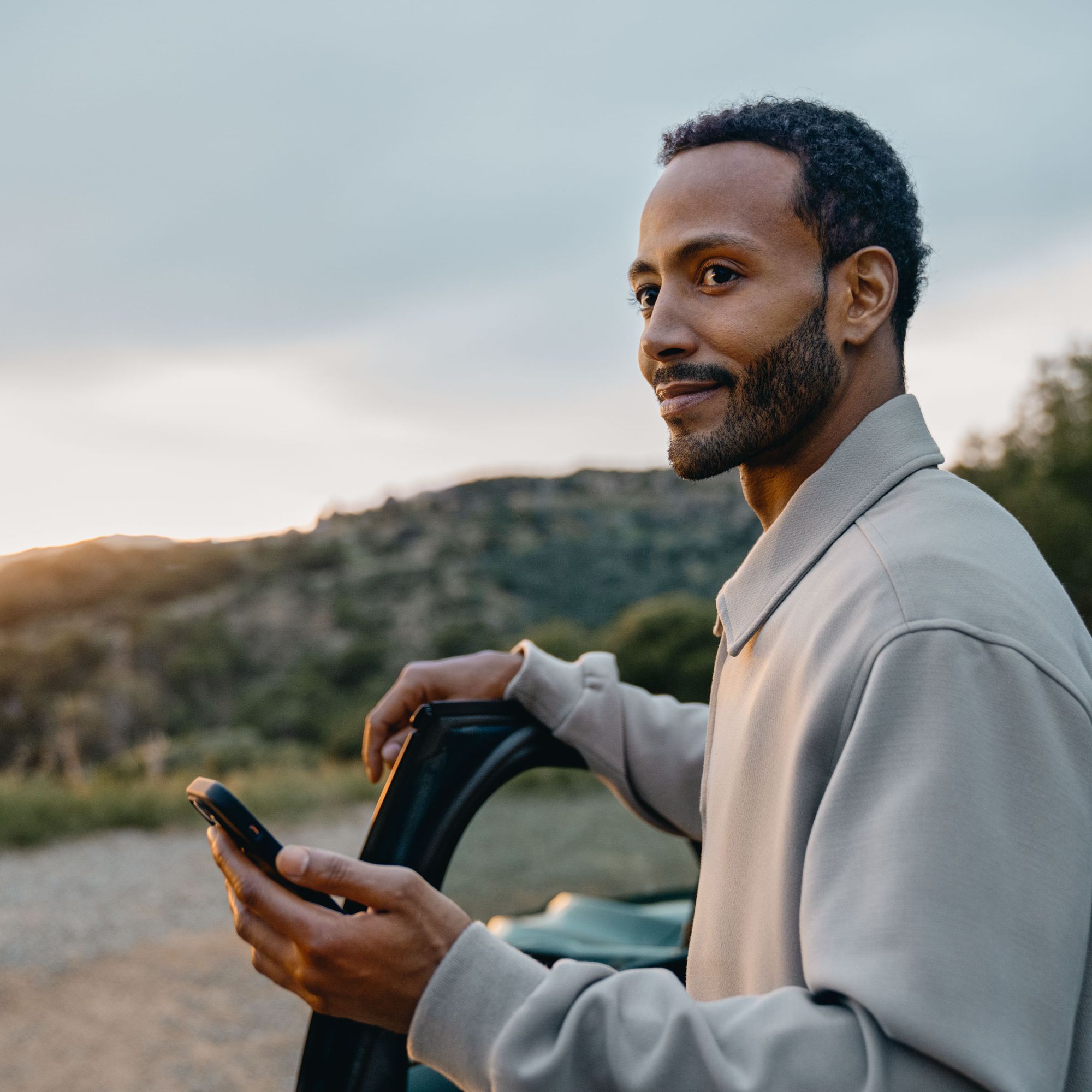 Ein junger Mann steht an der offenen Autotür und hält ein Smartphone in der Hand. Er blickt seitlich lächelnd in die Ferne, während im Hintergrund eine hügelige Landschaft bei Sonnenuntergang zu sehen ist.