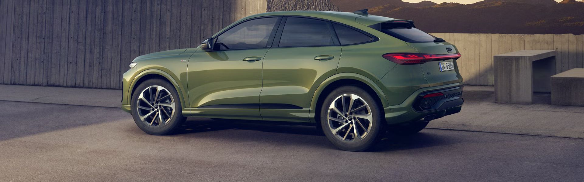Ein grüner Q5 Sportback in Seitenansicht steht auf einem Parkplatz vor einem urbanen Haus in bergiger Umgebung.