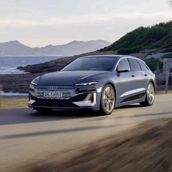 Der S6 Avant e-tron aus schräger Frontansicht, wie er eine Straße an der Küste entlangfährt. Im Hintergrund sind Berge zu sehen. 