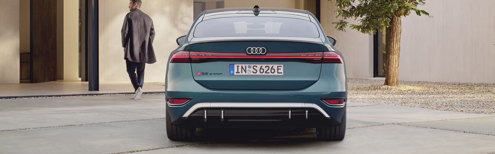 Der S6 Sportback e-tron im Heckprofil. Das Fahrzeug ist dem Eingangsbereich eines modernen Gebäudes zugewandt. Vor dem Auto befindet sich ein junger Mann, der auf das Gebäude zuläuft.