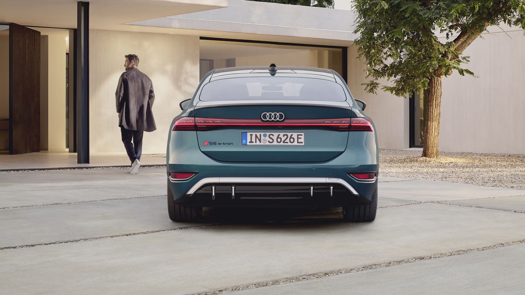 Der S6 Sportback e-tron im Heckprofil. Das Fahrzeug ist dem Eingangsbereich eines modernen Gebäudes zugewandt. Vor dem Auto befindet sich ein junger Mann, der auf das Gebäude zuläuft.