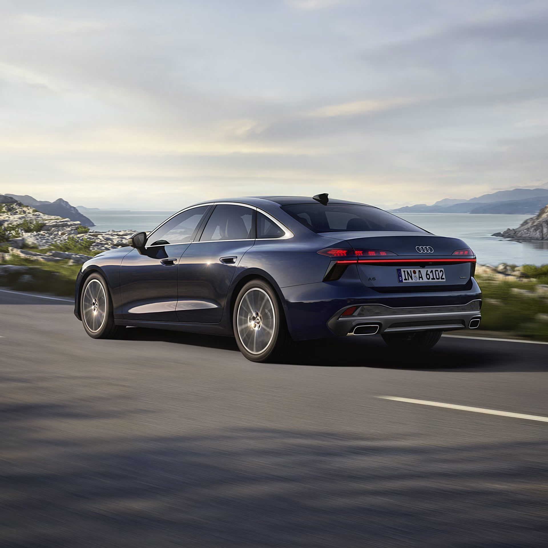 Die A6 Limousine in einer schrägen Heckperspektive, während sie auf einer kurvigen Straße entlang eines Fjords fährt. 