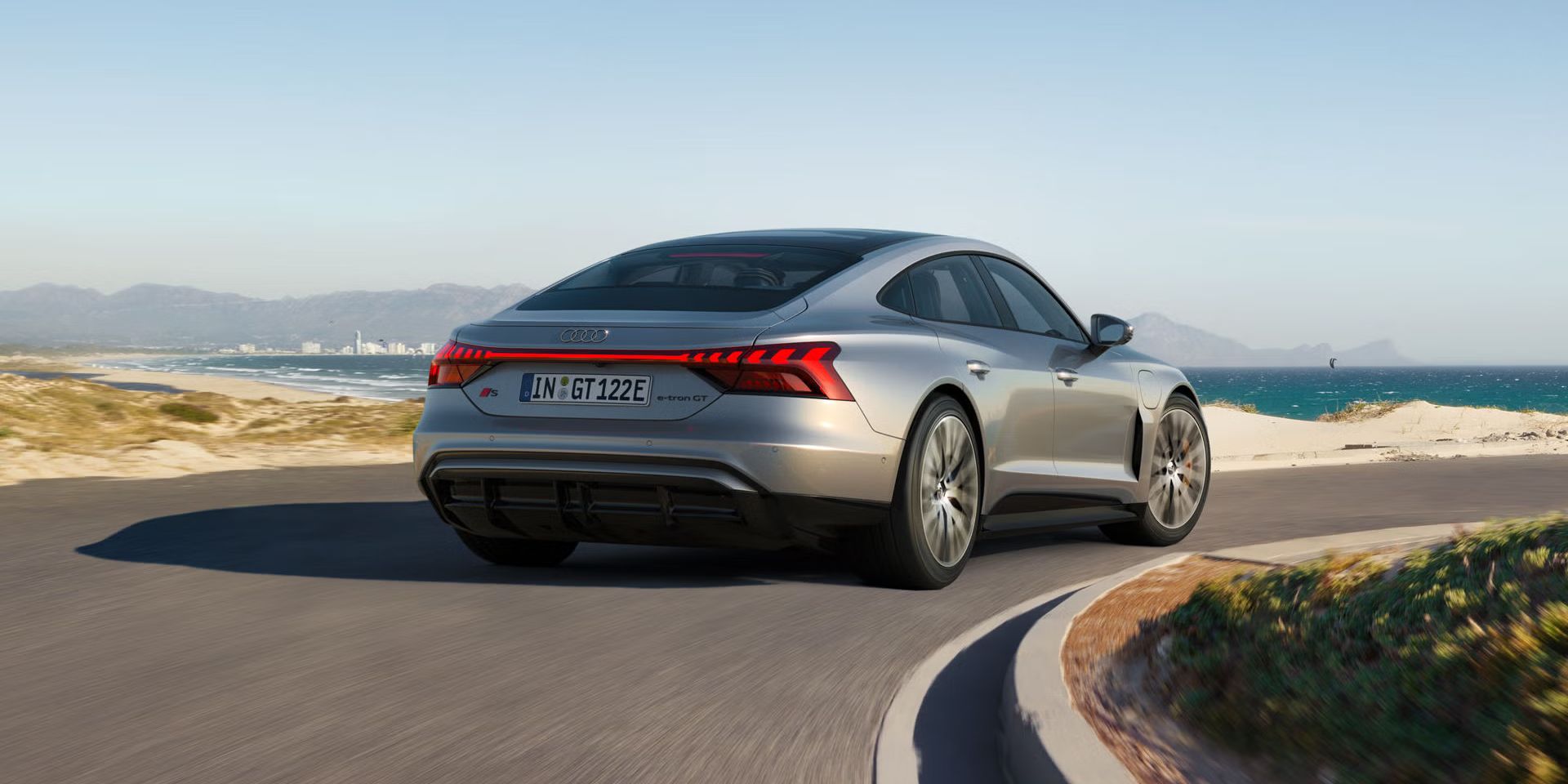 Ein silberner Audi S e-tron GT fährt auf einer kurvigen Küstenstraße mit Blick auf das Meer und eine Stadt im Hintergrund.