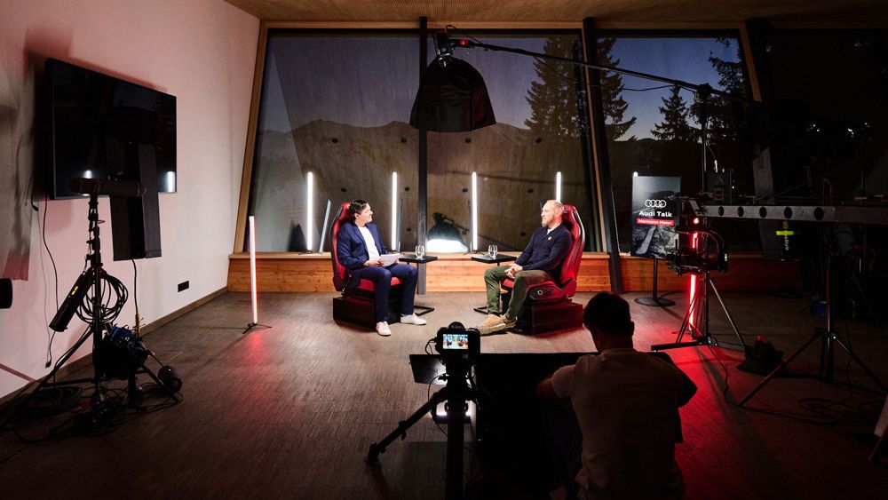 Hermann Maier und ein weiterer Mann sitzen in bequemen Sesseln in einem modern eingerichteten Raum und führen ein Interview. Das Setup beinhaltet Kameras, Lichter und technische Ausrüstung.