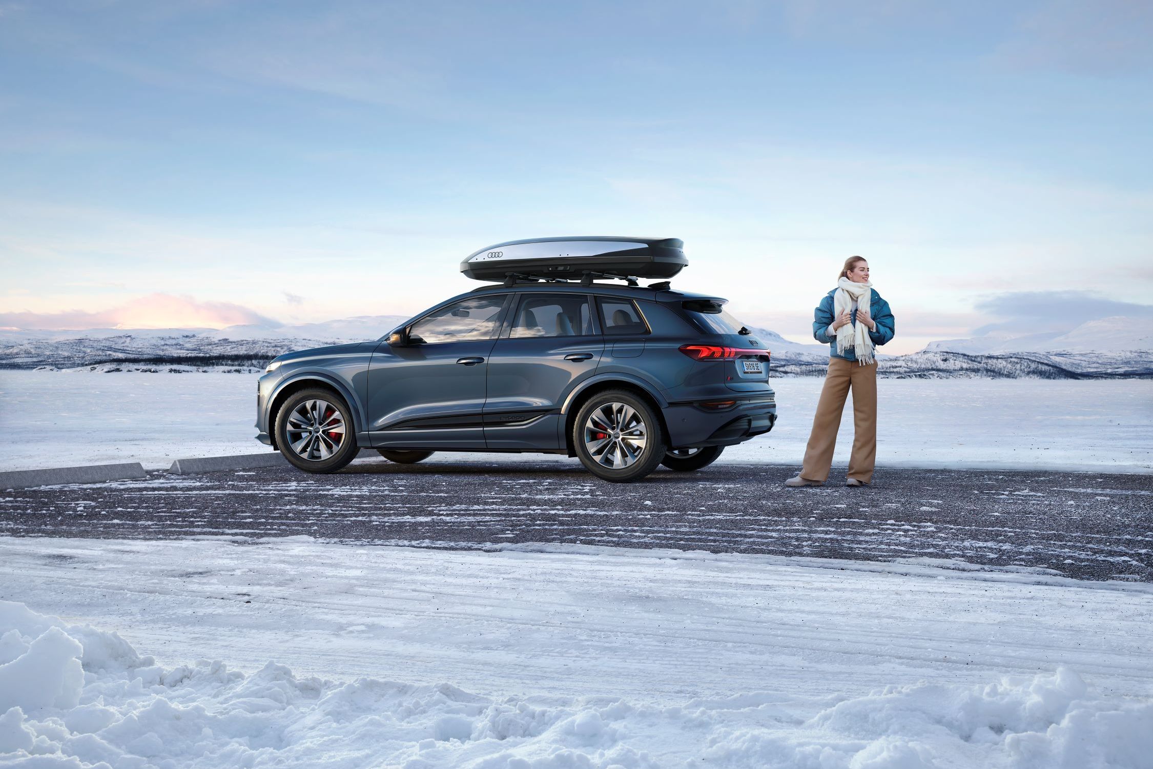 Ein Audi Q6 mit Dachbox parkt an einer winterlichen Straße, eine Frau steht davor