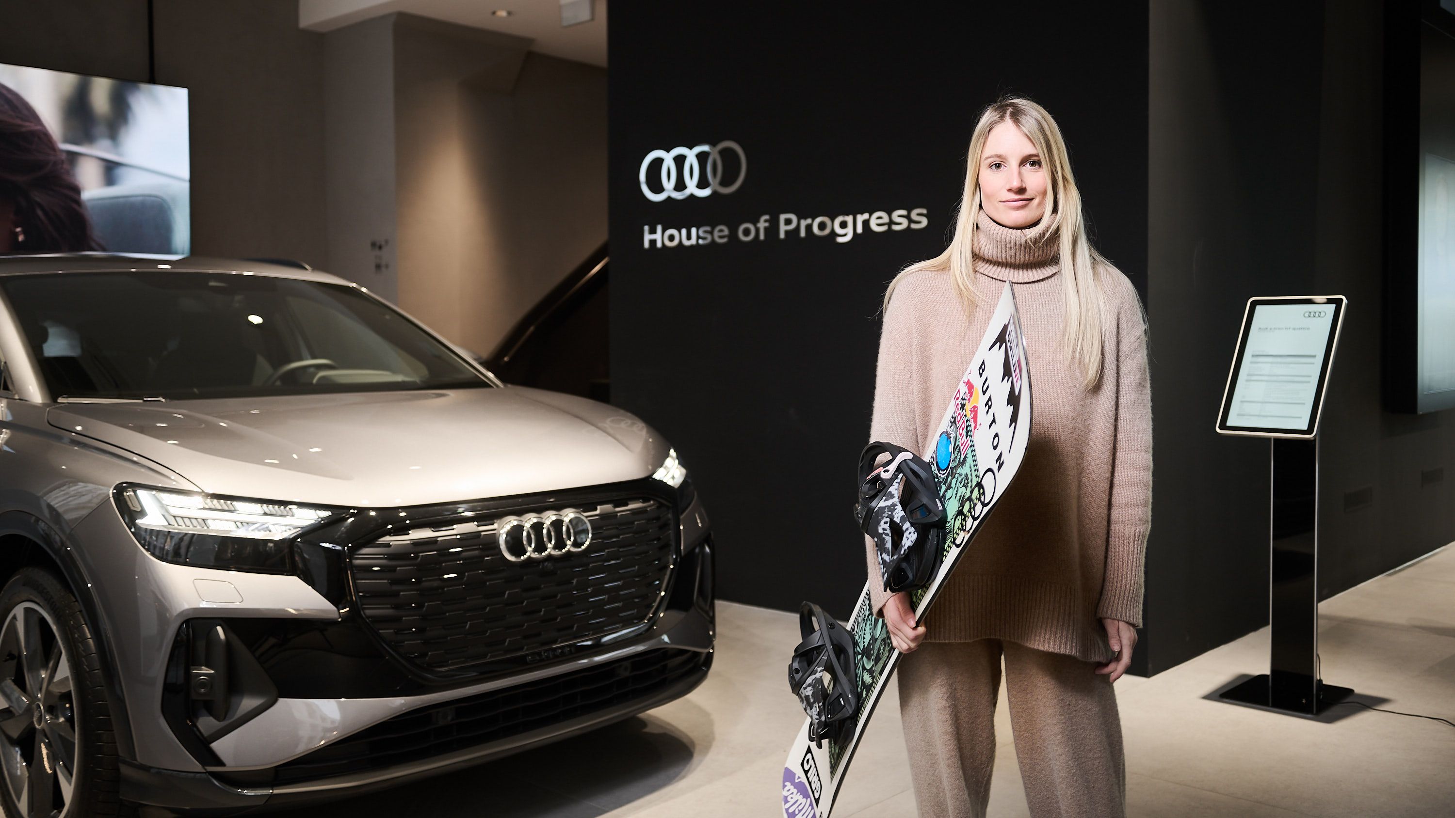 Anna Gasser in beigefarbener Kleidung steht vor einem Audi und hält ein Snowboard. Im Hintergrund ist ein Schild mit dem Audi-Logo und dem Text "House of Progress" zu sehen.