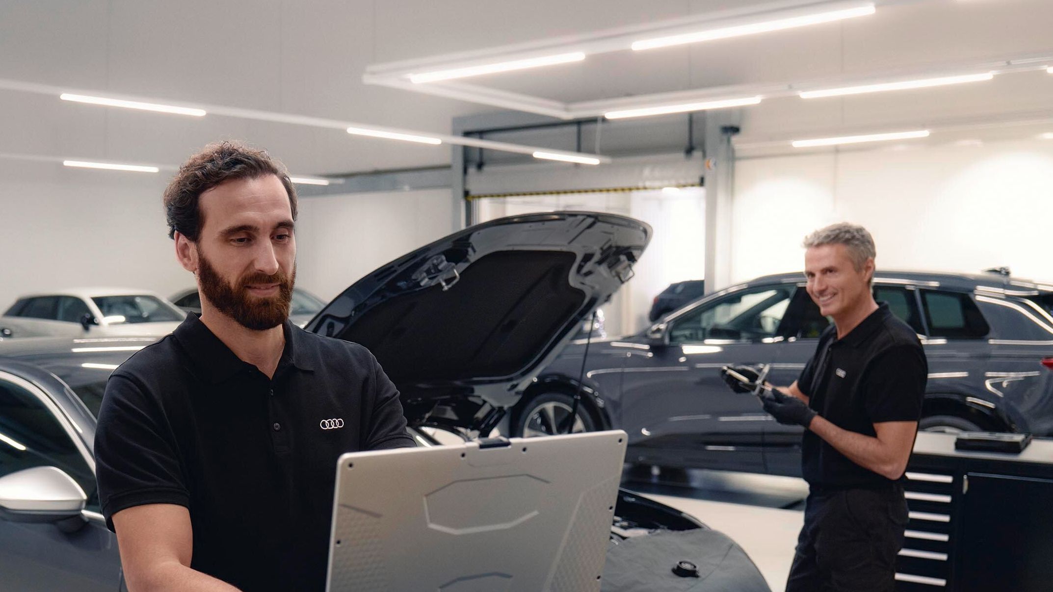 Zwei Audi Service Techniker in einer Werkstatt unterhalten sich. Einer von beiden blickt dabei auf seinen Laptop.