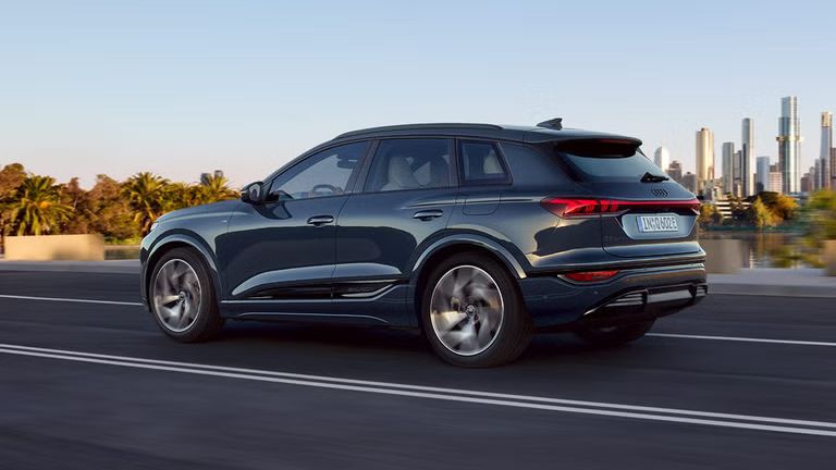 Seitenprofil des Q6 SUV e-tron, welcher auf einer Straße an einem Fluss fährt. Im Hintergrund ist die Skyline einer Stadt zu sehen.