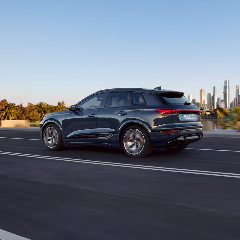 Seitenprofil des Q6 SUV e-tron, welcher auf einer Straße an einem Fluss fährt. Im Hintergrund ist die Skyline einer Stadt zu sehen.