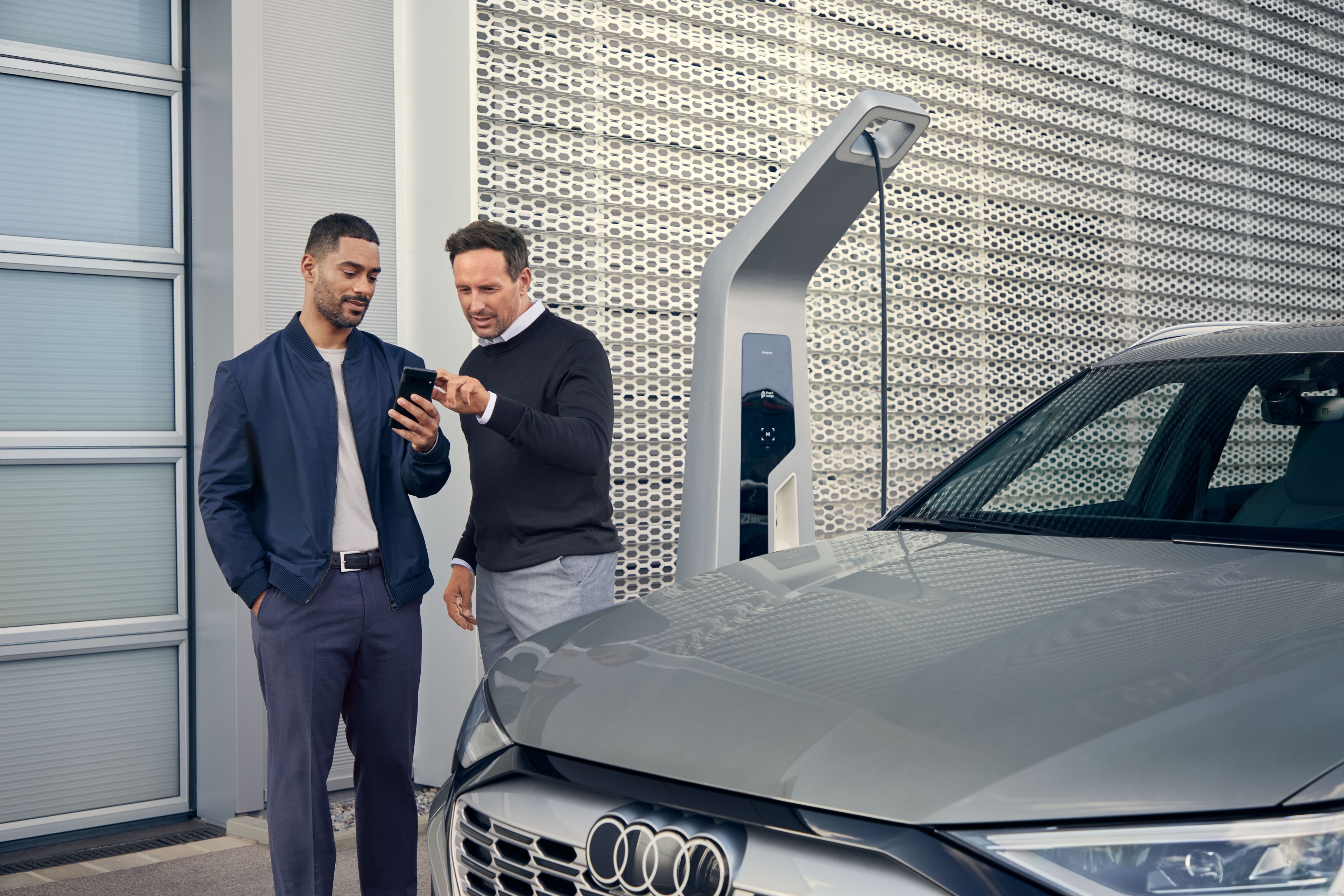 Ein Audi Service Mitarbeiter und ein Kunde blicken auf ein Smartphone, im Vordergrund steht ein Audi Elektroauto