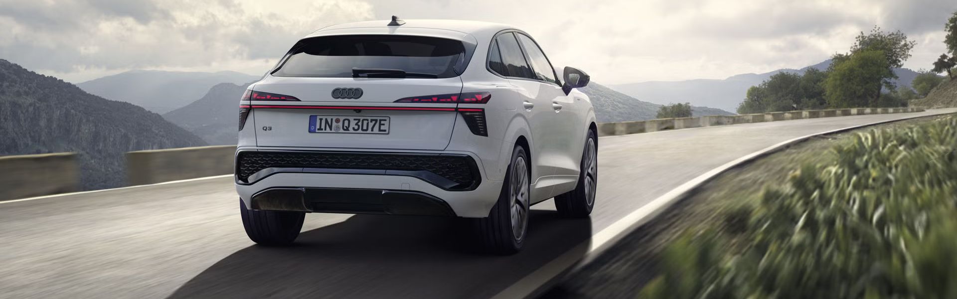 Ein weißer Q3 Sportback e-hybrid fährt auf einer Landstraße in einer bergigen Landschaft.	
