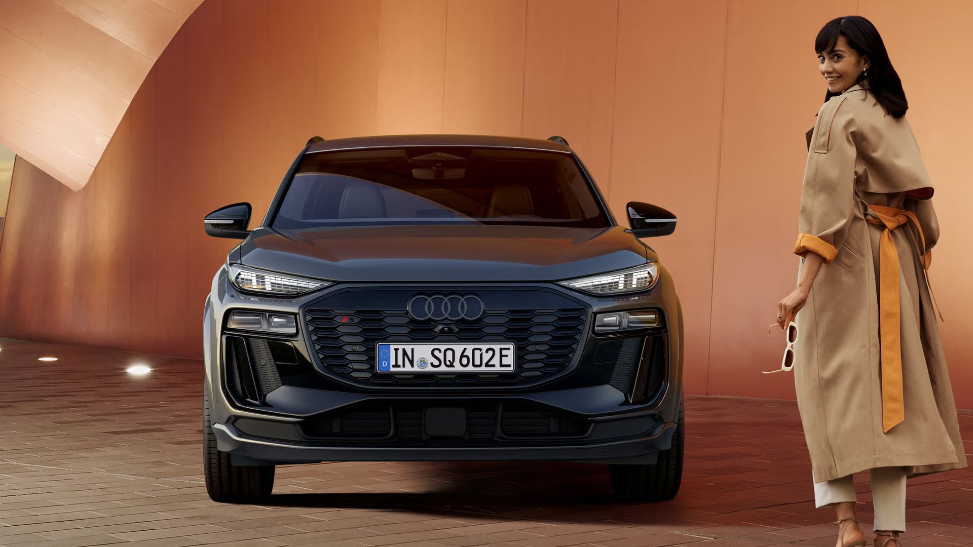 Frontprofil des SQ6 SUV e-tron, welcher vor einer orangenen, beleuchteten Wand steht. Auf der rechten Seite des Bildes geht eine junge Frau auf das Fahrzeug zu und schaut in die Richtung der Kamera zurück.