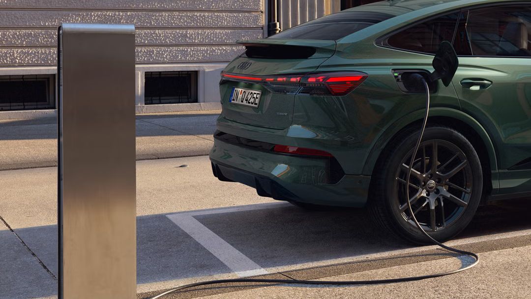 Blick auf das rechte Heck des Q4 Sportback e-tron, an einer Ladesäule parkend. Im Hintergrund ein modernes Gebäude. Das Kabel der Ladesäule ist mit dem Fahrzeug verbunden.