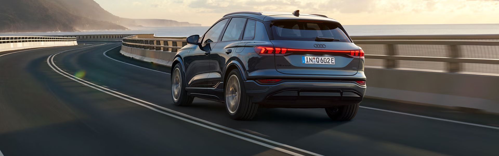 Schräge Heckaufnahme des Q6 SUV e-tron, welche über eine kurvige Straße am Meer fährt. Im Hintergrund sind Berge zu sehen.