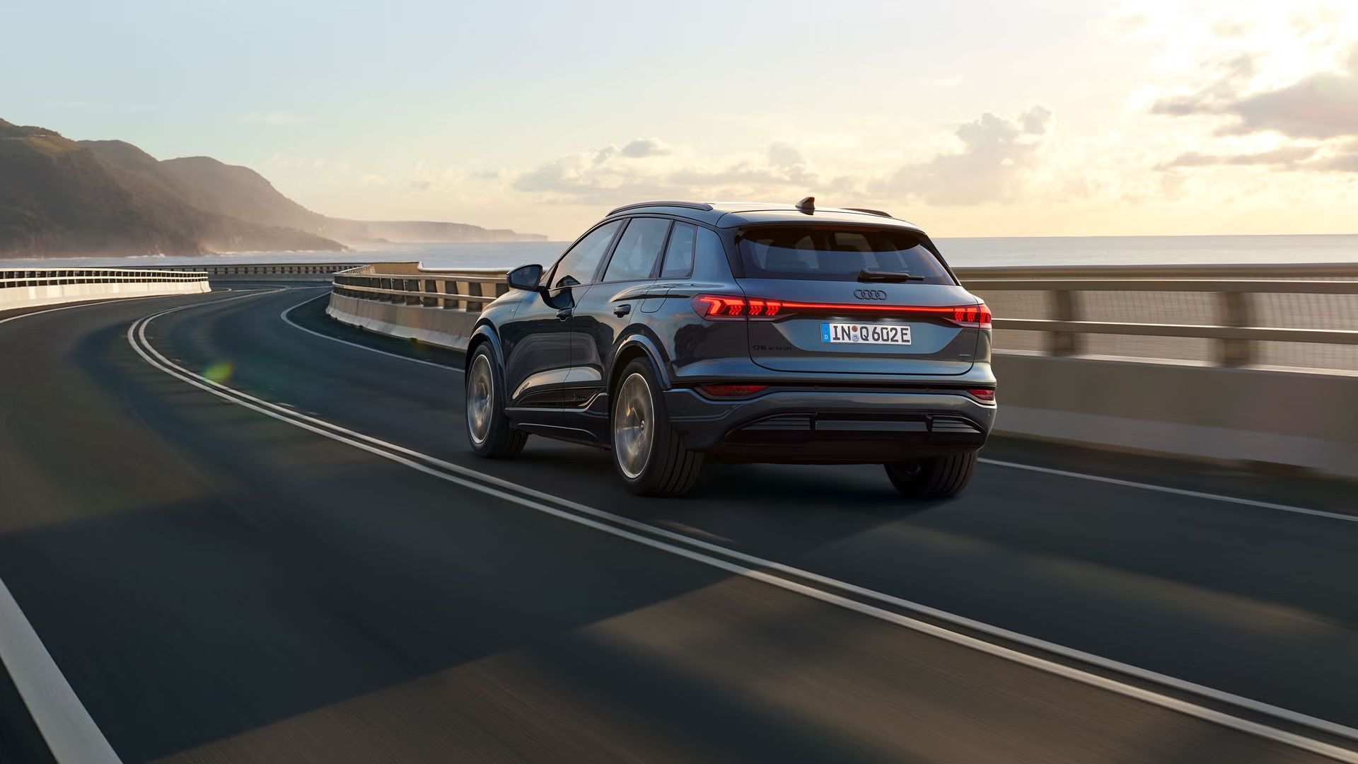 Schräge Heckaufnahme des Q6 SUV e-tron, welche über eine kurvige Straße am Meer fährt. Im Hintergrund sind Berge zu sehen.