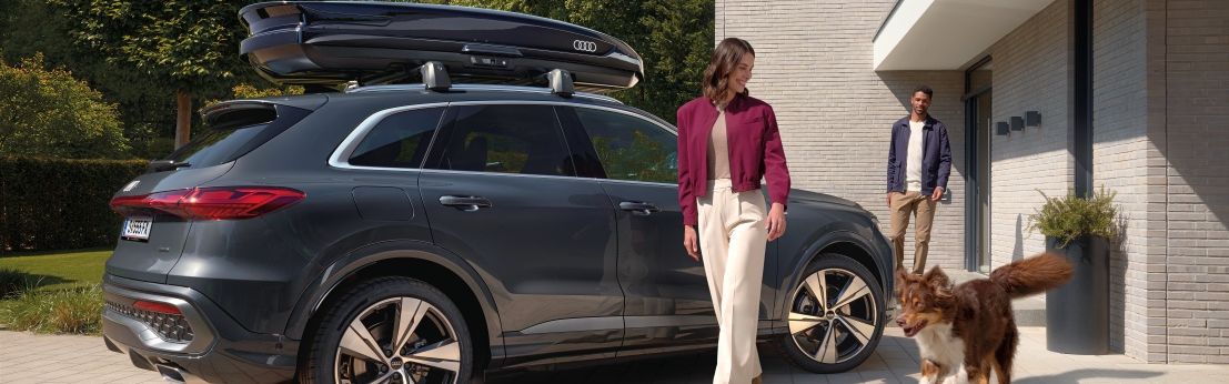 Ein Australian Shepard läuft freudig neben einer lächelnden Frau und einem parkenden Audi Q5 mit Dachbox umher während ein Mann vor dem Hauseingang hinter den beiden steht