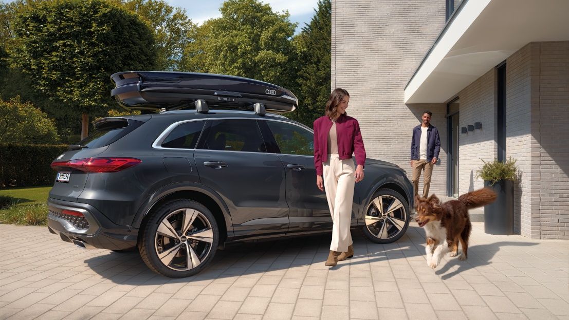 Ein Australian Shepard läuft freudig neben einer lächelnden Frau und einem parkenden Audi Q5 mit Dachbox umher während ein Mann vor dem Hauseingang hinter den beiden steht