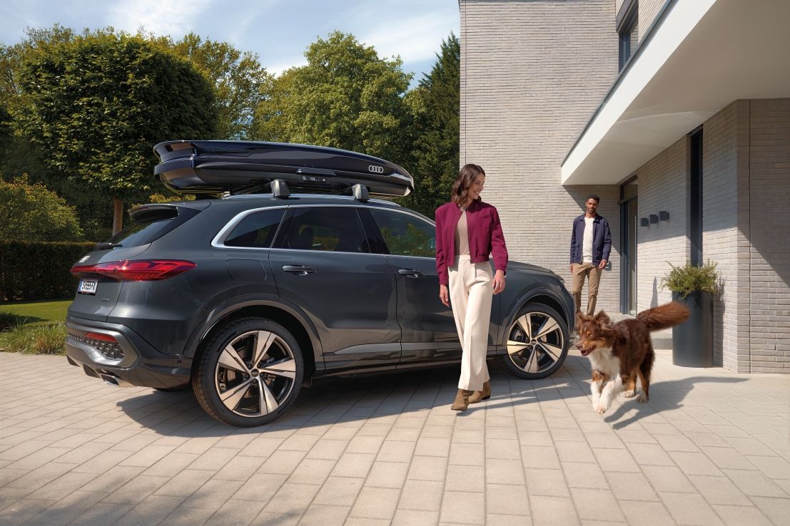 Ein Australian Shepard läuft freudig neben einer lächelnden Frau und einem parkenden Audi Q5 mit Dachbox umher während ein Mann vor dem Hauseingang hinter den beiden steht