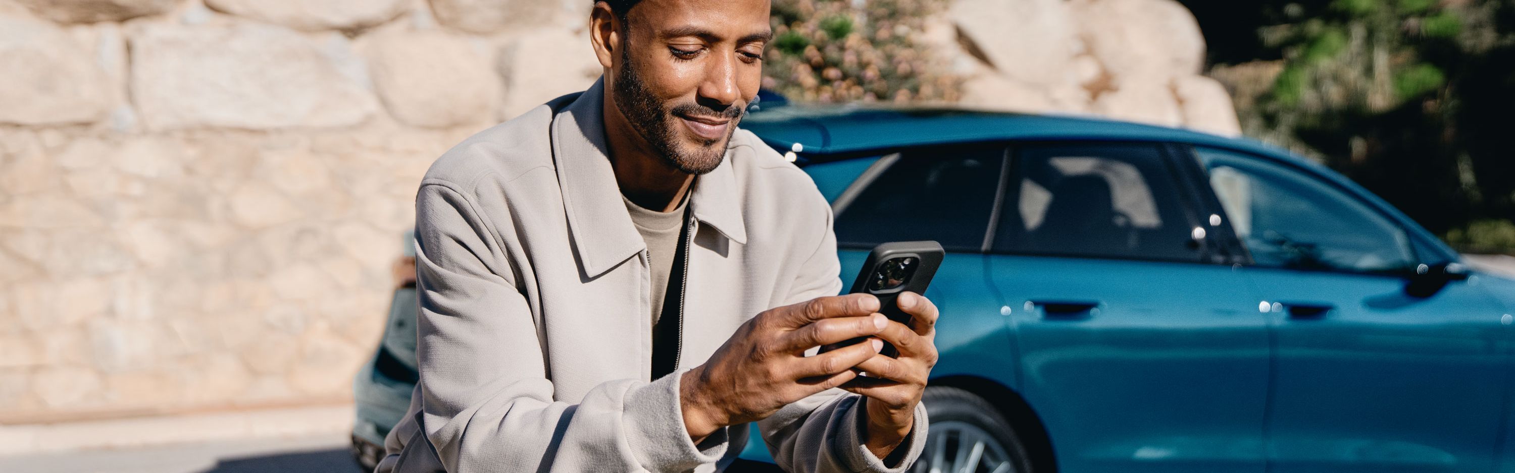 Ein Mann sitzt im Freien auf einer niedrigen Steinmauer und schaut lächelnd auf sein Smartphone. Hinter ihm stehen ein blauer Audi.