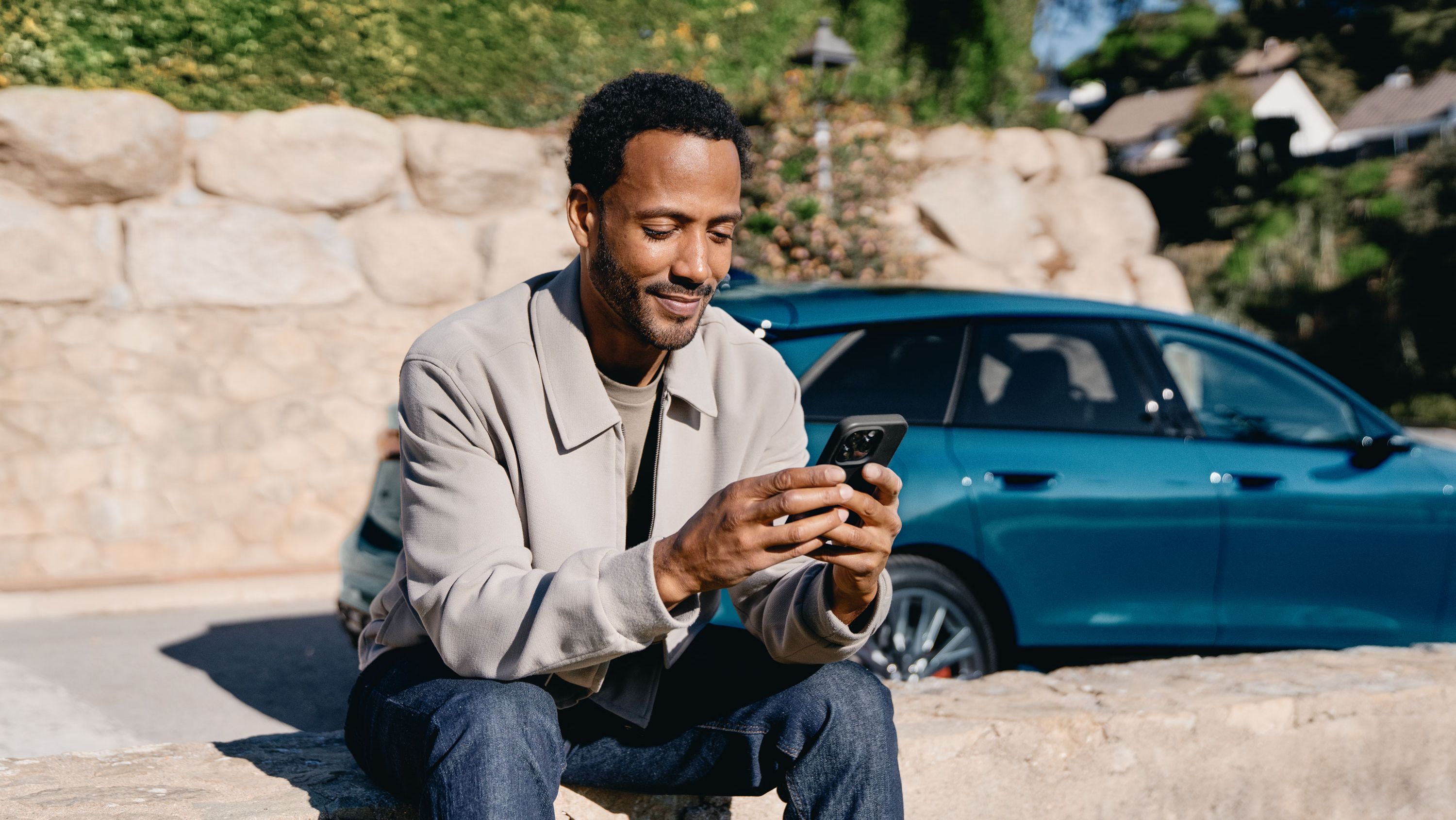 Ein Mann sitzt im Freien auf einer niedrigen Steinmauer und schaut lächelnd auf sein Smartphone. Hinter ihm stehen ein blauer Audi.