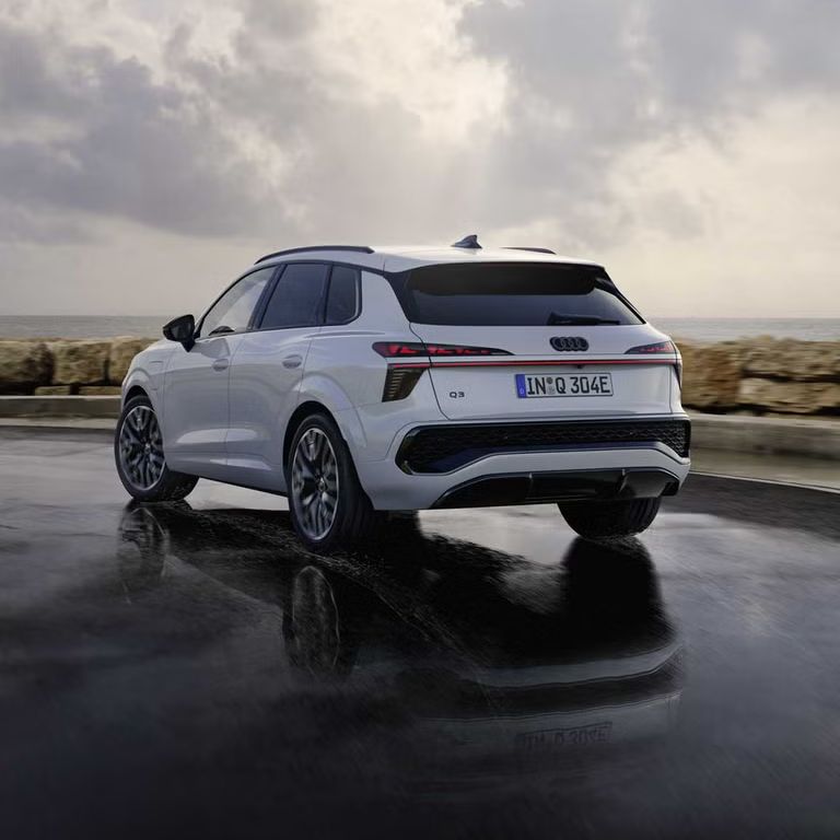 Ein weißer Q3 SUV e-hybrid von hinten fährt auf nasser Straße entlang der Küste.	