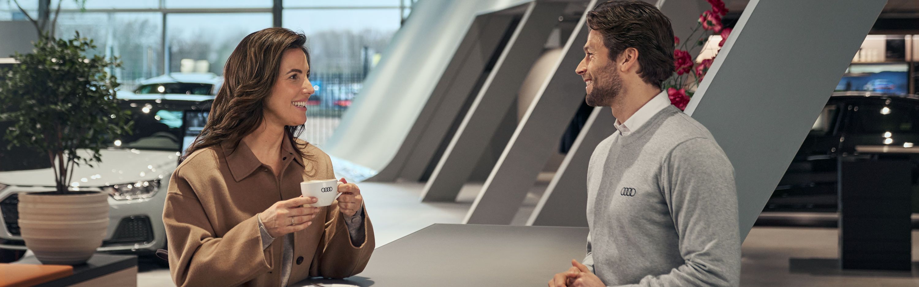 Ein Serviceberater im Gespräch mit einem Kunden in der Kaffeelounge; der Kunde hält eine Kaffeetasse in der Hand, im Hintergrund sind der Fahrzeugausstellungsbereich und der Kundenwartebereich zu sehen.
