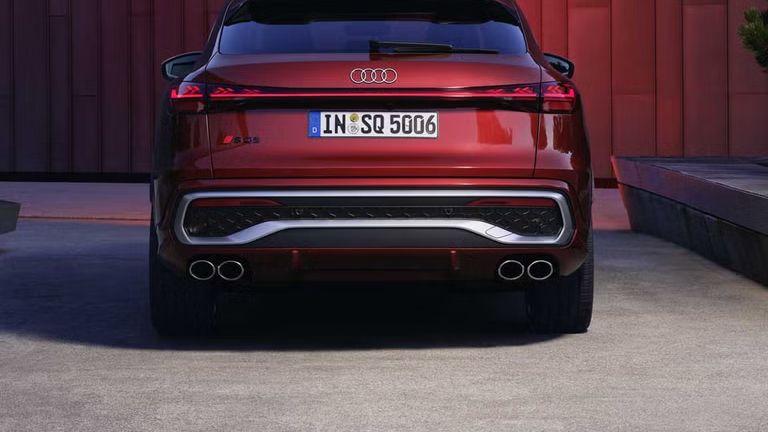 Heckprofil des roten SQ5 Sportback, welcher in einem Stadtpark vor einer Gebäudefassade steht und diese rot anleuchtet.