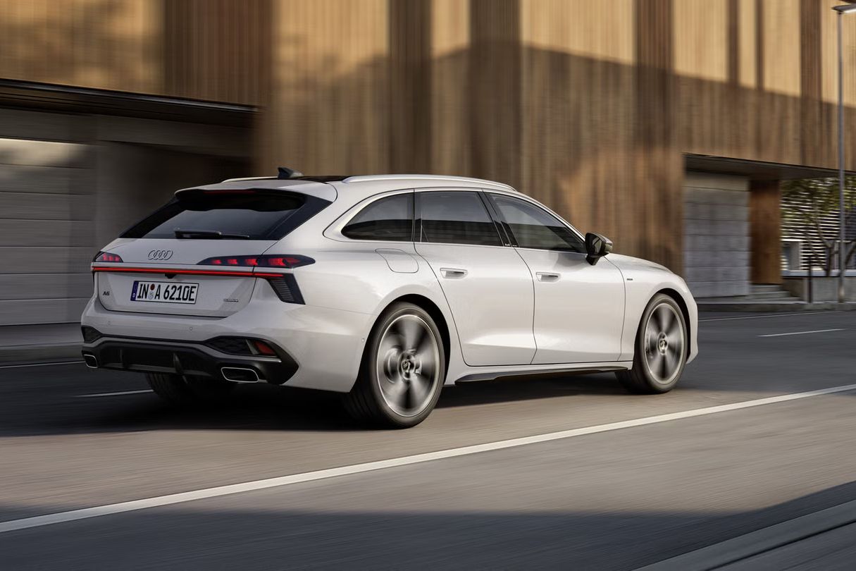 Blick von schräg hinten auf den A6 Avant e-hybrid, der auf einer Straße vor einem Gebäude entlangfährt.