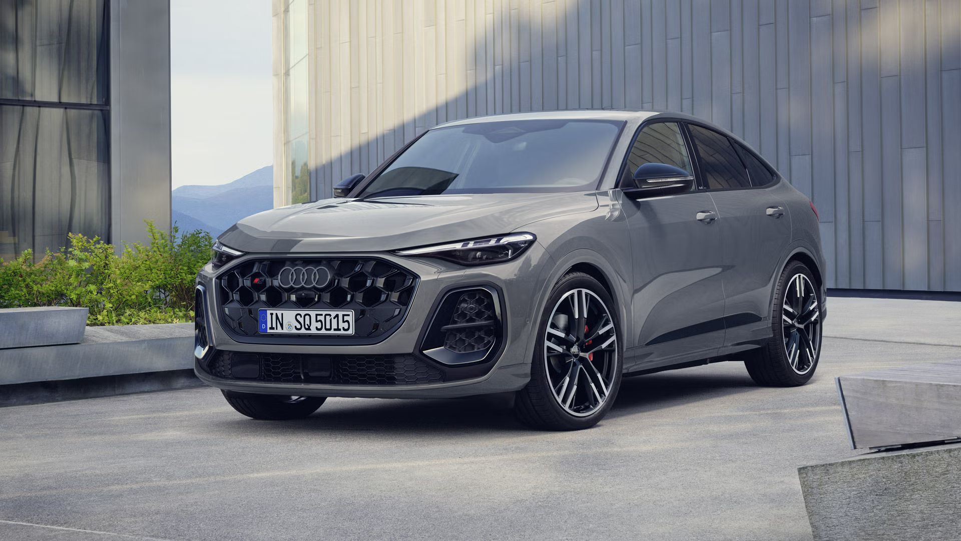 Der SQ5 Sportback steht im schrägen Frontprofil vor einem großen Gebäude. Das Fahrzeug ist Nardograu lackiert.