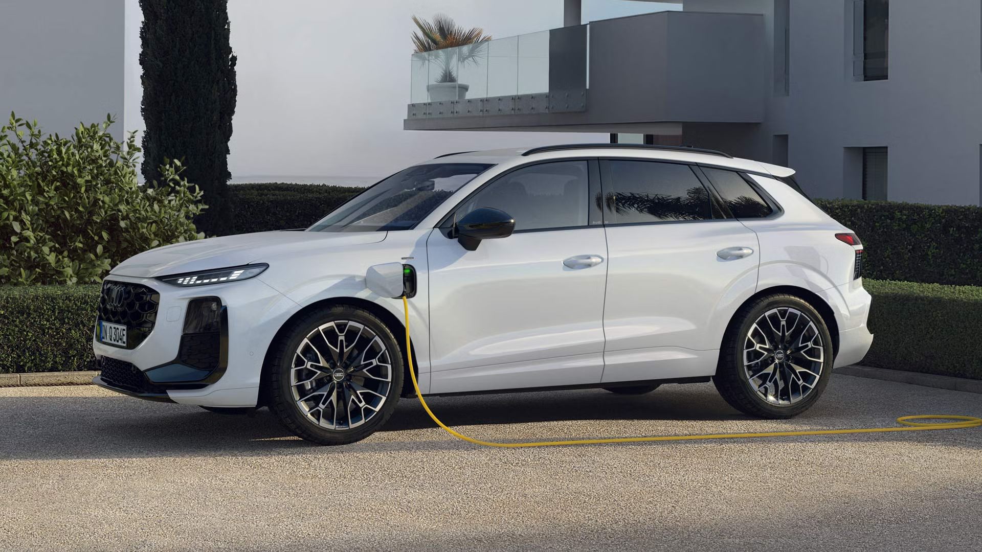Seitenprofil eines weißen Q3 SUV e-hybrid, der auf dem Parkplatz vor einem modernen Haus lädt.