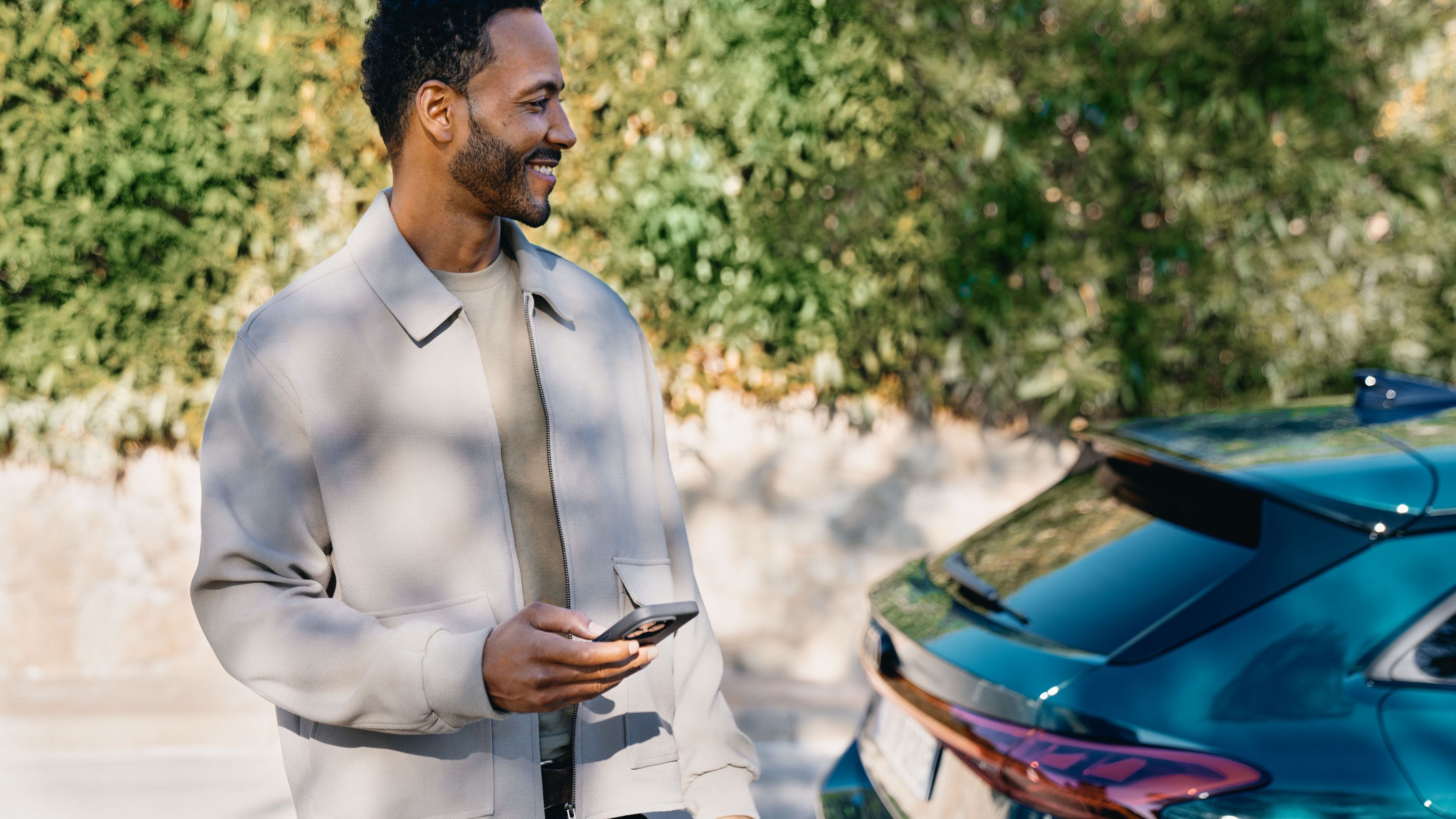 Ein lächelnder Mann steht im Freien vor einer grünen Hecke und hält ein Smartphone in der Hand. Er trägt eine hellbeige Jacke und schaut nach links aus dem Bild. Rechts im Vordergrund ist das Heck eines dunkelgrünen Audi zu sehen.