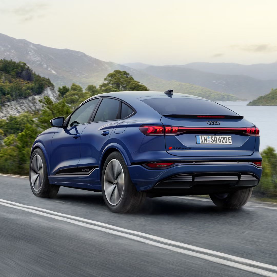 Schräge Heckaufnahme des SQ6 Sportback e-tron, welcher eine Straße an einem See in einer bergigen Landschaft entlangfährt.