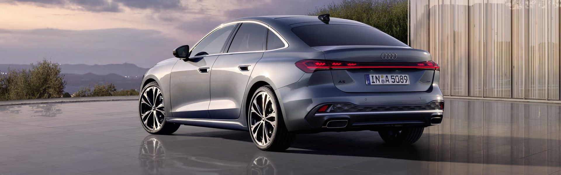 Die A5 Limousine im schrägen Heckprofil. Das Fahrzeug spiegelt sich auf dem glatten Untergrund. Im Hintergrund sind die Konturen von Bergen bei Dämmerung zu sehen.