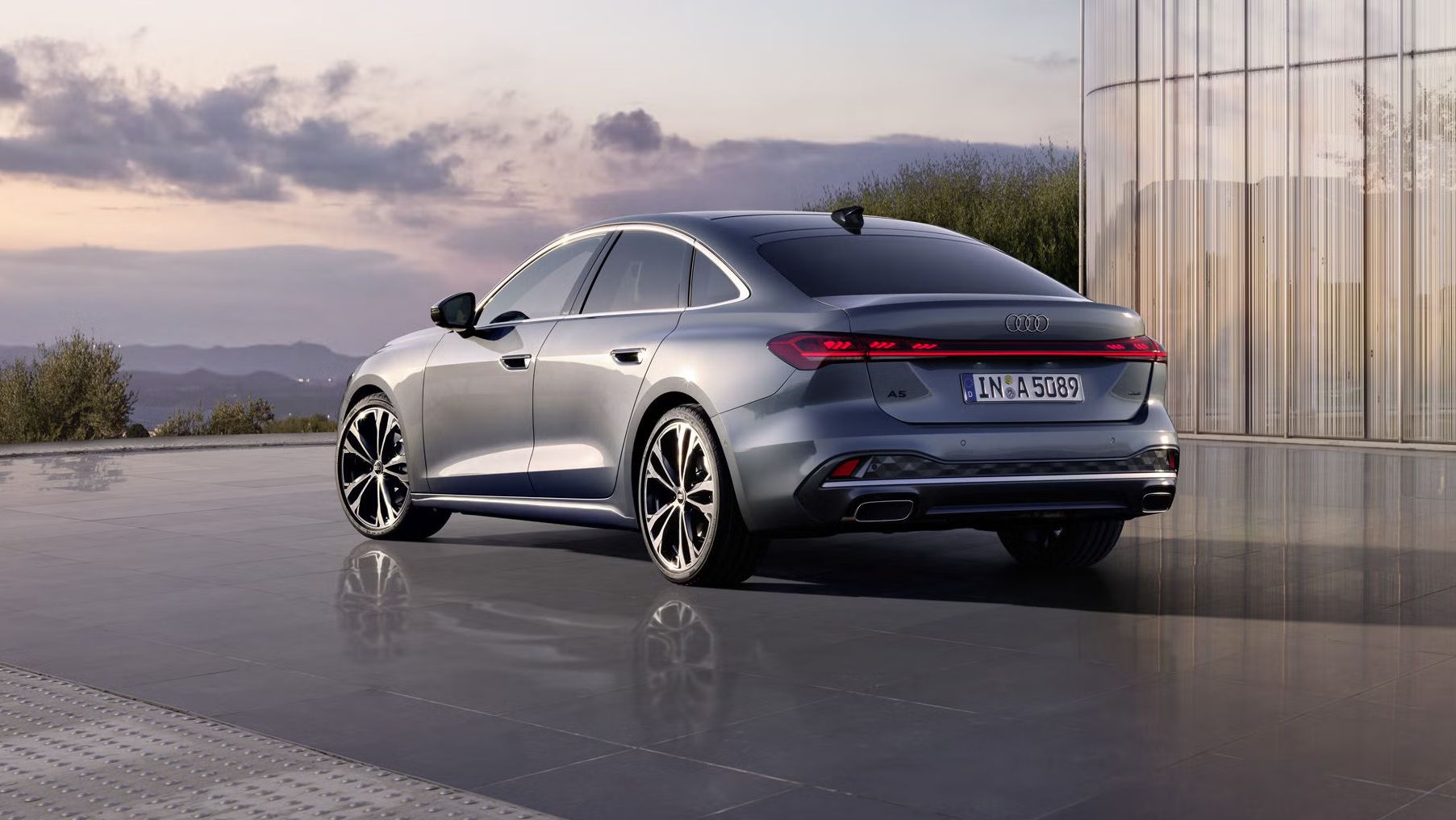 Die A5 Limousine im schrägen Heckprofil. Das Fahrzeug spiegelt sich auf dem glatten Untergrund. Im Hintergrund sind die Konturen von Bergen bei Dämmerung zu sehen.