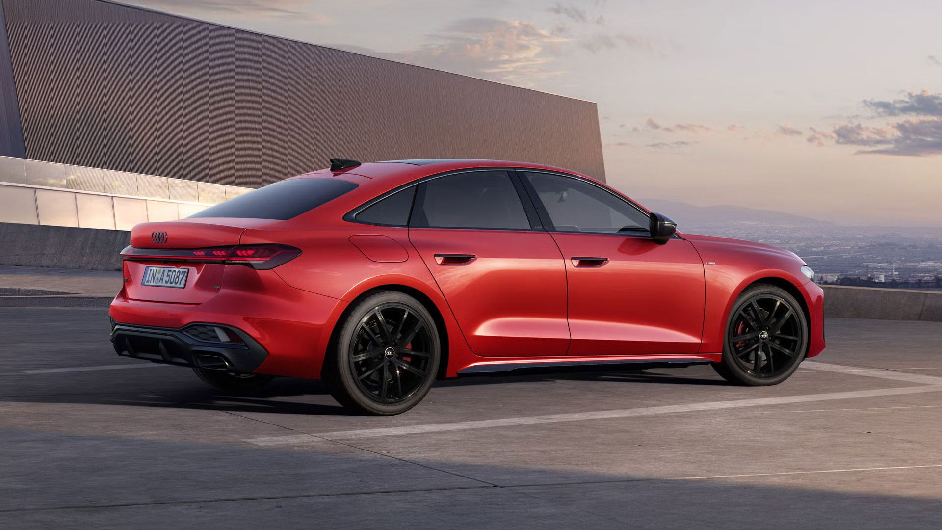 Die A5 Limousine im schrägen Seitenprofil. Das Fahrzeug ist misanorot lackiert und steht auf einem Parkdeck vor einem großen Gebäude.