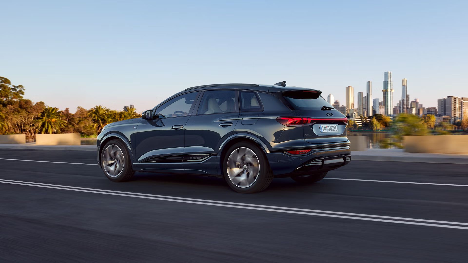 Seitenprofil des Q6 SUV e-tron, welcher auf einer Straße an einem Fluss fährt. Im Hintergrund ist die Skyline einer Großstadt zu sehen.